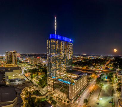 Nighttime urban skyline featuring a modern glass high-rise with blue crown lights and rooftop pool, city streets aglow and a full moon on the horizon.