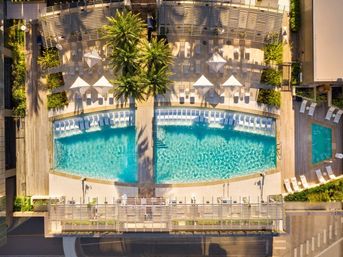 Aerial view of a sunny urban rooftop pool with turquoise water, palm trees, white loungers and umbrellas on a wooden sun deck.