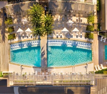 Aerial view of a sunny urban rooftop pool with turquoise water, palm trees, white loungers and umbrellas on a wooden sun deck.