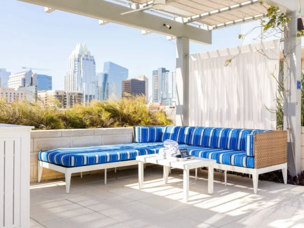 Sunlit urban rooftop lounge with blue-striped sectional sofa, white side tables under a pergola, ornamental grasses and a city skyline in the background