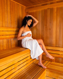 Relaxed woman wrapped in a white towel sitting barefoot on wooden benches inside a warm cedar‑paneled sauna, enjoying a peaceful spa moment.
