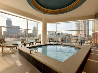 Sunlit indoor rooftop spa pool with panoramic city skyline views, striped lounge chairs around a square soaking pool and modern high-rise backdrop.