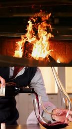 Roaring fireplace above a close-up of red wine being poured into an elegant curved glass decanter, evoking a cozy, warm home dining atmosphere.
