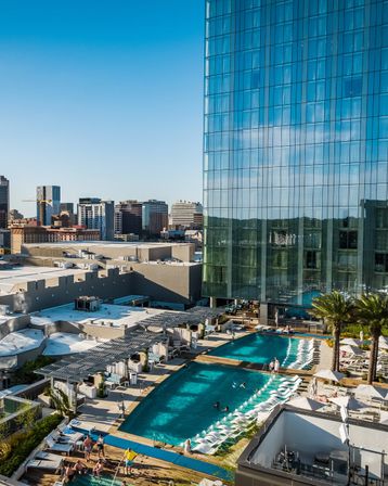 Rooftop resort-style pool and sun deck with lounge chairs and palm trees beside a reflective glass skyscraper, overlooking a sunny downtown skyline.