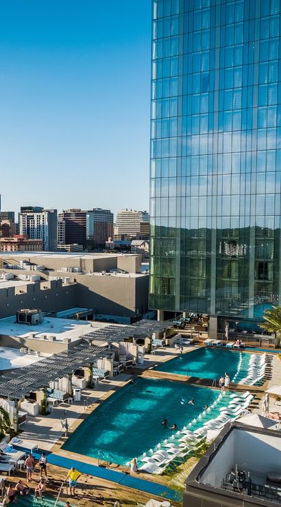 Rooftop resort-style pool and sun deck with lounge chairs and palm trees beside a reflective glass skyscraper, overlooking a sunny downtown skyline.