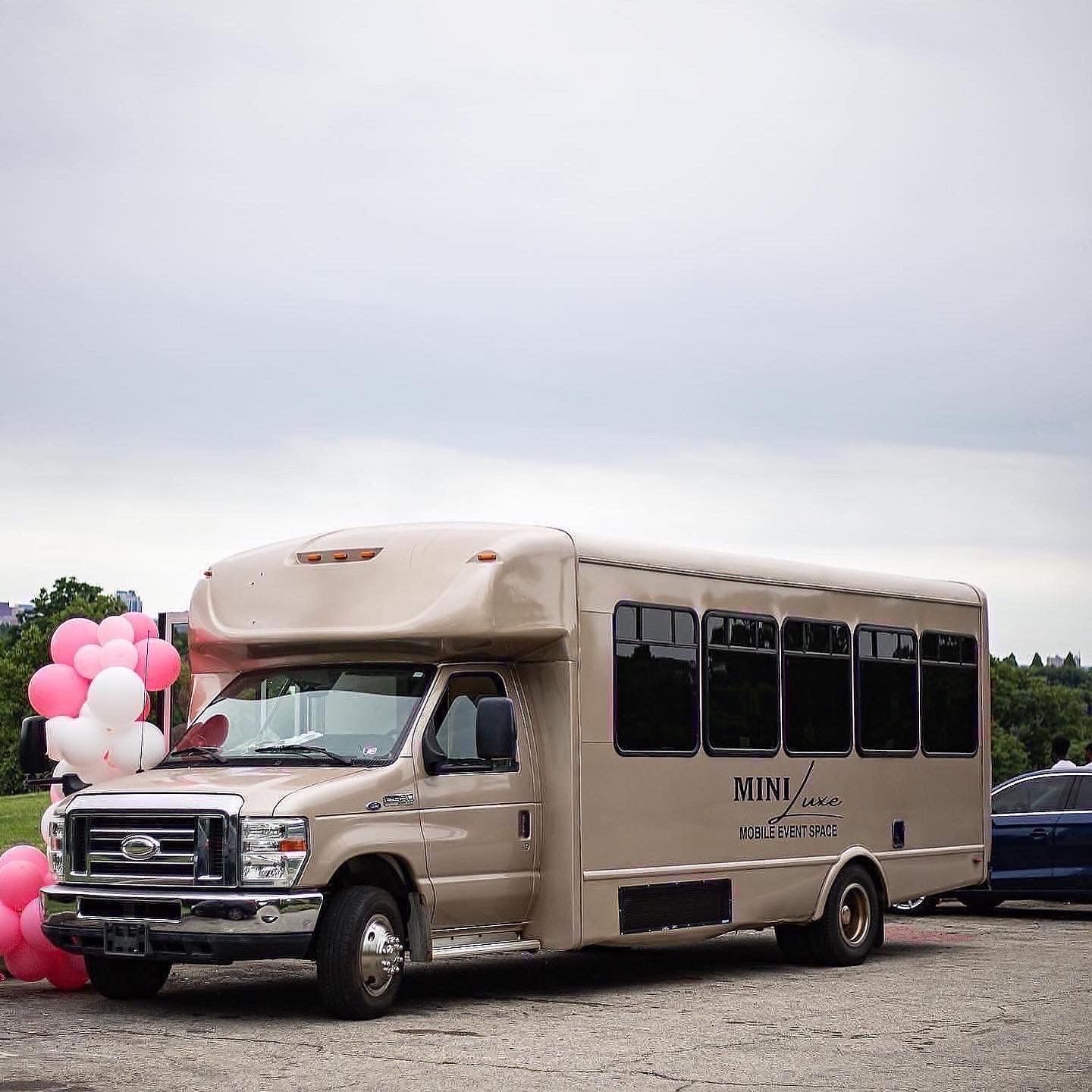 Beige mobile event shuttle parked in a lot with pink and white balloons tied to the front, overcast sky and tree-lined background
