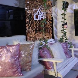 White wooden swing with ivy-wrapped chains and faux roses, styled with fluffy white and pink rose-gold sequin pillows in front of a copper sequin backdrop and geometric white wall — glamorous indoor lounge photo-op