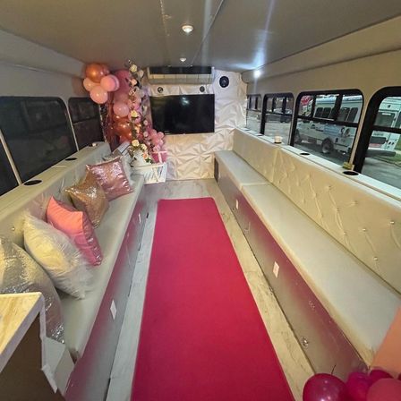 Luxury pink-themed party bus interior with long tufted white benches, sequined pillows, floral arrangement, pink balloons, a bright pink aisle runner and wall-mounted TV.