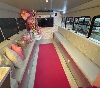 Luxury pink-themed party bus interior with long tufted white benches, sequined pillows, floral arrangement, pink balloons, a bright pink aisle runner and wall-mounted TV.