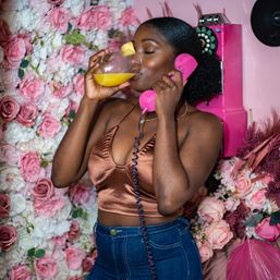 Woman in a bronze satin crop top sipping orange juice with a pineapple garnish while holding a pink retro handset in front of a pink rose floral wall