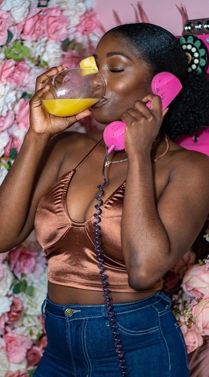 Woman in a bronze satin crop top sipping orange juice with a pineapple garnish while holding a pink retro handset in front of a pink rose floral wall
