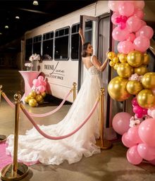 Bride in a lace wedding gown with long train stepping into a white mobile event shuttle at an indoor venue, framed by pink-and-gold balloon arch, pink velvet stanchions and a floral cocktail table.