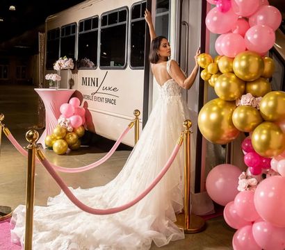 Bride in a lace wedding gown with long train stepping into a white mobile event shuttle at an indoor venue, framed by pink-and-gold balloon arch, pink velvet stanchions and a floral cocktail table.