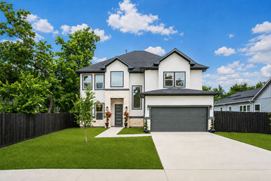 Modern two-story suburban home with white stucco and dark trim, attached two-car garage, concrete driveway, manicured green lawn, small front tree and tall leafy trees under a bright blue sky with puffy clouds.