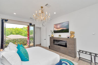 Bright airy modern bedroom with a large bed and teal accent pillows, sparkling crystal chandelier, wall-mounted TV above a wood fireplace console, and open French doors to a balcony with leafy green views.