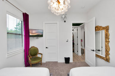 Bright modern bedroom with white walls and carpet, twin beds, a bold purple curtain, green vintage armchair, wall-mounted TV, gold ornate mirror, bubble chandelier and an open door into a hallway.