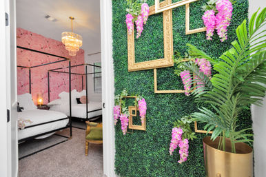 Entry view of a stylish bedroom with two black-frame canopy beds and white bedding, pink flamingo wallpaper, a crystal chandelier, and a decorative boxwood accent wall with gold picture frames, hanging pink blossoms and a potted palm.