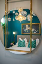 Reflection in a round mirror showing a stylish bedroom with teal accent wall, crystal chandelier and hanging disco balls, framed leopard painting, upholstered headboard and white bed topped with green patterned pillows.