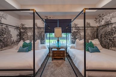 Cozy symmetrical guest bedroom with two black four-poster twin beds dressed in white linens, teal patterned and blue accent pillows, an ornate wooden nightstand topped by a blue glass lamp in front of a window, and black-and-white tropical wallpaper mural.