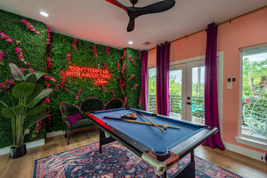 Vibrant indoor game room with a blue pool table, cues and racked balls, tufted velvet sofa against a lush faux green wall with pink blooms and a glowing neon quote sign, magenta curtains, hardwood floor and balcony view of trees.