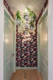 Decorated narrow indoor hallway with dark floral wallpaper, mint-green doors, hanging disco balls, cascading white flowers and greenery, and a neon 'Happy Birthday' sign.