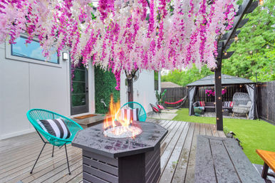 Backyard patio with a pergola draped in pink and white hanging wisteria, a lit fire pit on a wooden deck, teal chairs with striped pillows, gazebo lounge, hammock, and string lights over artificial turf.