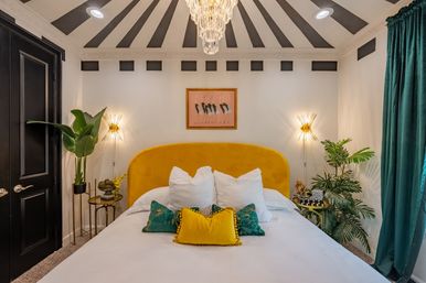 Stylish modern glam bedroom with mustard yellow velvet headboard, white bedding and colorful throw pillows, teal curtains, tropical plants, gold sunburst wall sconces and a crystal chandelier under a black-and-white radial ceiling.