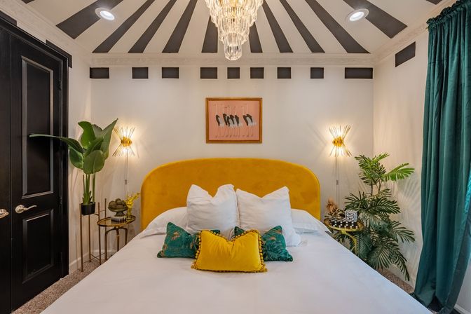 Stylish modern glam bedroom with mustard yellow velvet headboard, white bedding and colorful throw pillows, teal curtains, tropical plants, gold sunburst wall sconces and a crystal chandelier under a black-and-white radial ceiling.
