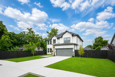 Sunny suburban two-story white home with gray two-car garage, long concrete driveway, manicured green lawn, young trees and a black wood fence under a bright blue sky with fluffy clouds.
