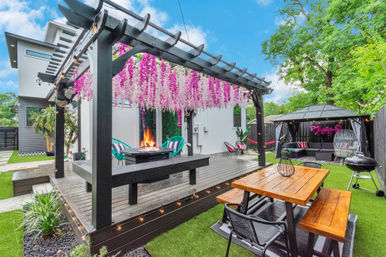 Modern backyard patio with a black pergola draped in pink hanging wisteria, central fire pit on a wooden deck, picnic table, gazebo lounge, string lights and a charcoal grill.