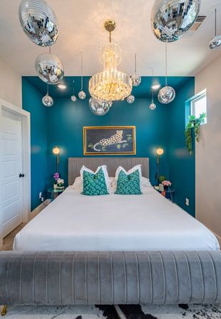 Modern glam teal bedroom with king bed and velvet gray bench, crystal chandelier and hanging disco balls, patterned teal accent pillows and framed leopard artwork.