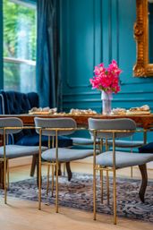 Luxury teal dining room with gold-accented gray velvet chairs around a wooden table, bright pink flower centerpiece, ornate gold mirror and garden-view window