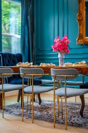 Luxury teal dining room with gold-accented gray velvet chairs around a wooden table, bright pink flower centerpiece, ornate gold mirror and garden-view window