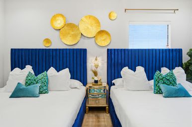 Bright modern twin bedroom with vertical blue velvet headboards, crisp white bedding, teal patterned throw pillows, gold circular wall sculptures and a mirrored gold nightstand
