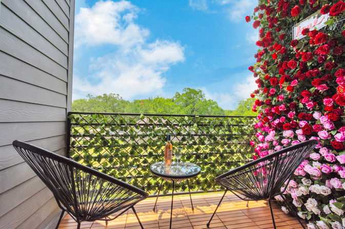 Cozy sunlit balcony with two black woven chairs and a small glass table holding a bottle of rosé and a wine glass, ivy-covered railing and vibrant red‑and‑pink rose wall overlooking green treetops and blue sky.