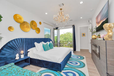 Bright modern luxury bedroom with navy velvet scalloped headboard, white linens, teal accent pillows, gold circular wall art, crystal chandelier and balcony overlooking treetops