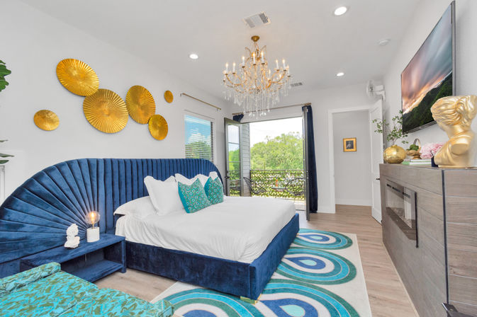Bright modern luxury bedroom with navy velvet scalloped headboard, white linens, teal accent pillows, gold circular wall art, crystal chandelier and balcony overlooking treetops