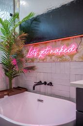 Modern bathroom with a white freestanding tub, matte black wall faucet, tropical palm plants and a pink neon sign reading "Lets get naked" over patterned tile.