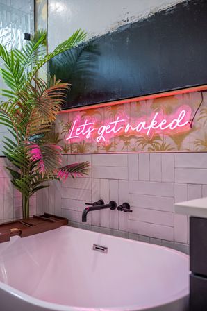 Modern bathroom with a white freestanding tub, matte black wall faucet, tropical palm plants and a pink neon sign reading "Lets get naked" over patterned tile.