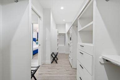 Bright modern walk-in closet with white built-in shelves and drawers, light wood-look flooring, recessed ceiling lights, luggage stand, and a glimpse of a blue upholstered bed in the adjacent bedroom.