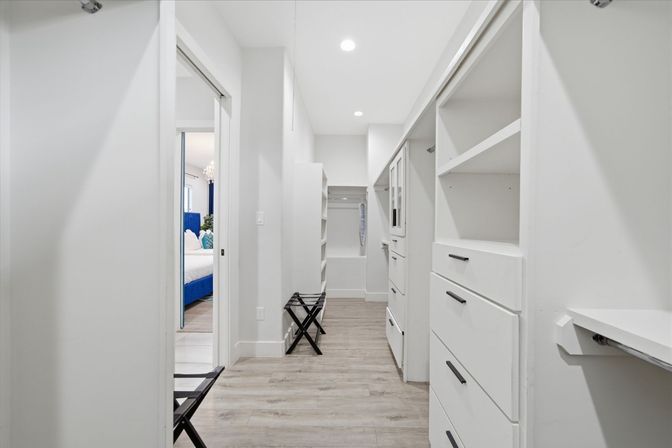 Bright modern walk-in closet with white built-in shelves and drawers, light wood-look flooring, recessed ceiling lights, luggage stand, and a glimpse of a blue upholstered bed in the adjacent bedroom.