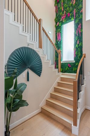 Bright modern entryway with light wood stairs, black metal balusters and oak handrail, teal decorative wall fan, potted tropical plant, and a lush vertical green wall with pink flowers framing a frosted window.