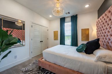 Stylish modern bedroom interior with a pink tufted velvet headboard, white linens, teal floor-length curtains, crystal chandelier, gold wall accents, zebra rug and potted plant.