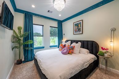 Bright bedroom with king bed, teal crown molding and curtains, crystal chandelier, colorful accent pillows, indoor plant and tree-lined window view.