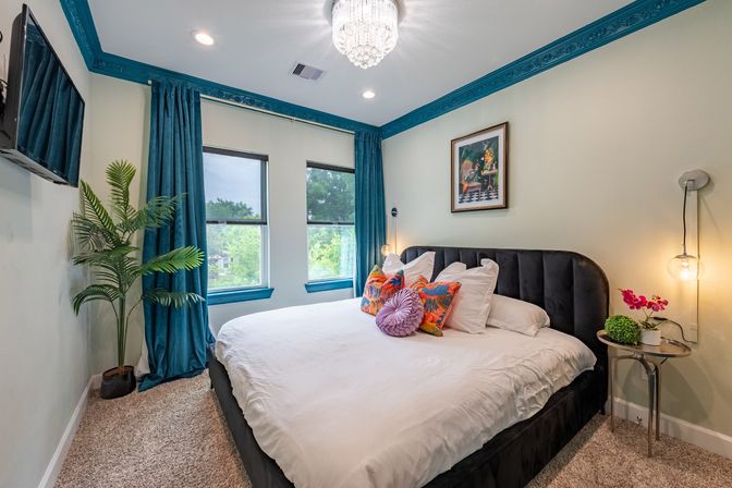 Bright bedroom with king bed, teal crown molding and curtains, crystal chandelier, colorful accent pillows, indoor plant and tree-lined window view.