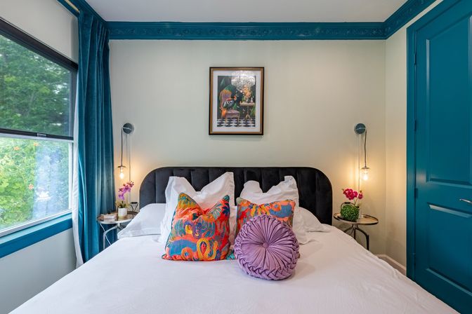 Bright modern bedroom with white bedding, black velvet headboard, teal crown molding and door, large window with teal curtains showing green trees, colorful patterned throw pillows and a round purple accent pillow, hanging bedside lights