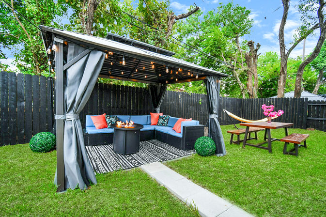 Cozy backyard pergola with gray curtains and string lights, blue corner sectional around a lit fire pit on a patterned rug, next to a wooden picnic table with hammock and pink flowers, green lawn and tall privacy fence
