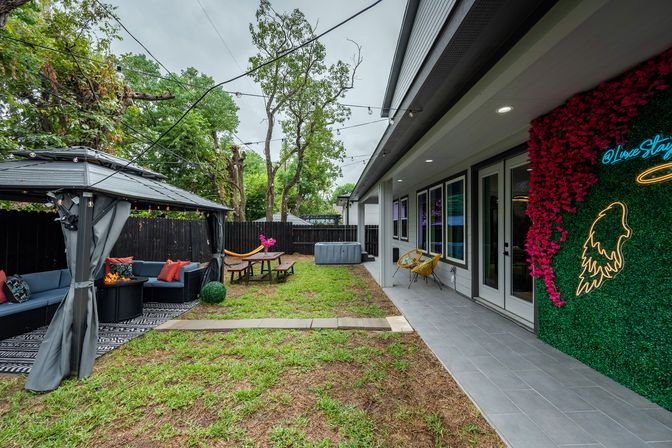 Modern suburban backyard patio with covered gazebo lounge, string lights, outdoor sectional and fire pit, picnic table with hammock, hot tub, tiled porch and decorative green wall with neon art.