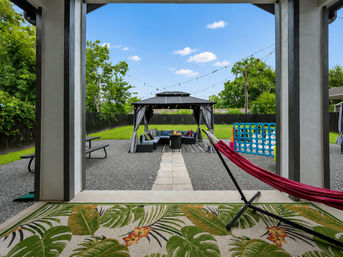 Inviting suburban backyard patio featuring a covered gazebo lounge with sectional seating around a fire table under string lights, a red hammock in the foreground, grassy lawn, picnic table and a large blue outdoor Connect Four game.