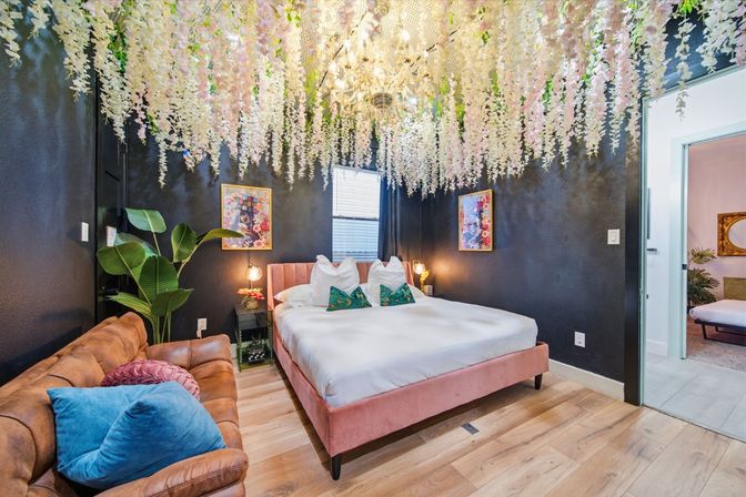 Dreamy boutique-style bedroom with cascading wisteria ceiling, crystal chandelier, pink velvet bed, black accent walls, leather sofa, tropical plant and wood plank flooring